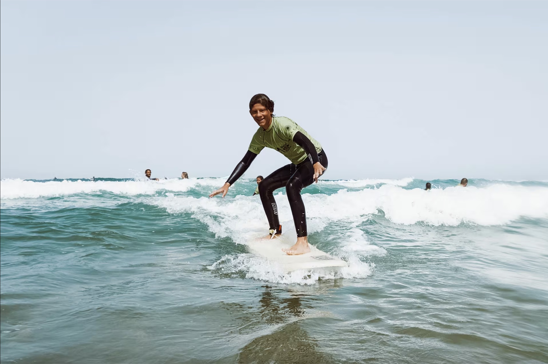 A smiling student standing up on a soft-top surfboard in the whitewater