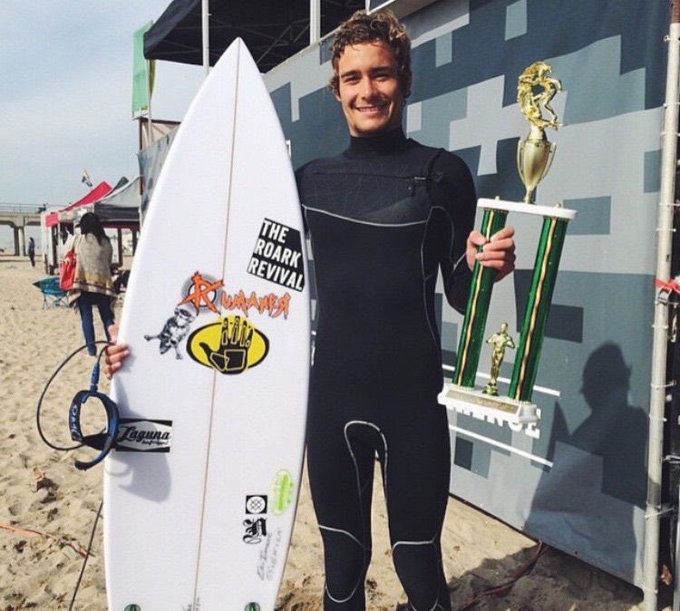 A young Lucas Taub in his wetsuit holding his surfboard and a contest trophy